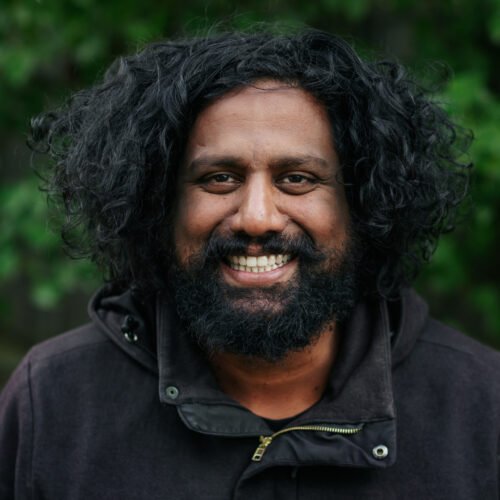 A dark skinned man with a beard and fluffy hair looking at the camera smiling.